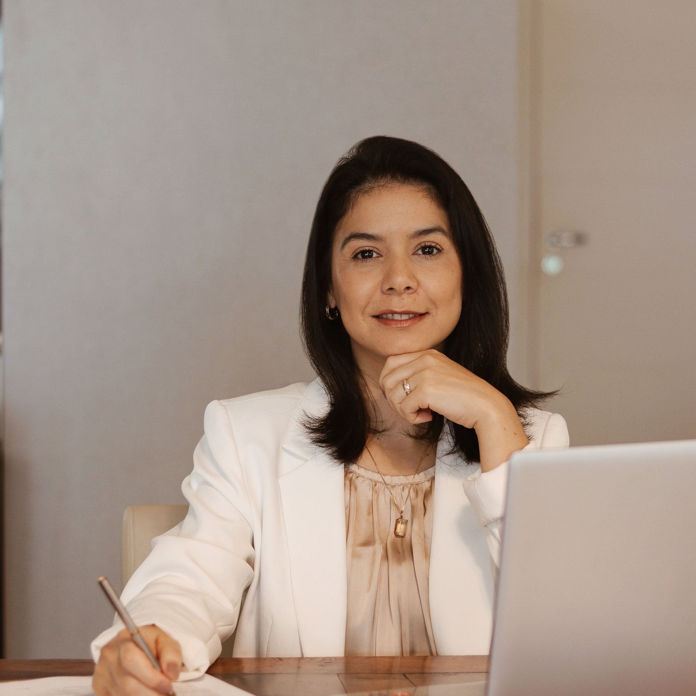 A woman in a white blazer sits at a desk with a laptop and notepad, pen in hand. She looks confident and professional, conveying focus and warmth.