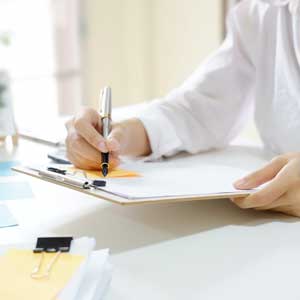 A person in a white shirt writes on a clipboard with a pen. The desk holds paper clips, documents, and notes, creating a focused and organized workspace.
