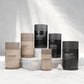 Six cylindrical supplement containers in shades of brown and black are arranged on a stone surface. Labels display ingredients like Cur cumin and Magnesium. The tone is minimal and modern.