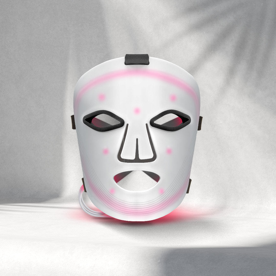 A white LED therapy mask with pink light accents is displayed against a textured Gray background. The mask has eye, nose, and mouth cutouts, conveying a futuristic feel.