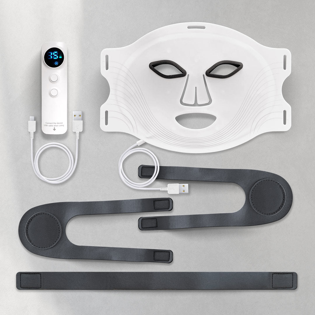 An LED skincare mask set with a white face mask featuring eye and mouth cutouts, a control device, USB cables, and adjustable black straps on a Gray surface.