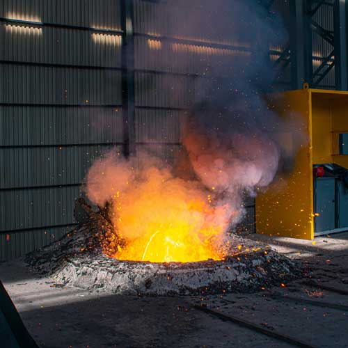 Molten metal glows with bright orange and yellow flames, casting sparks, in an industrial setting. Smoky haze rises against corrugated metal walls.