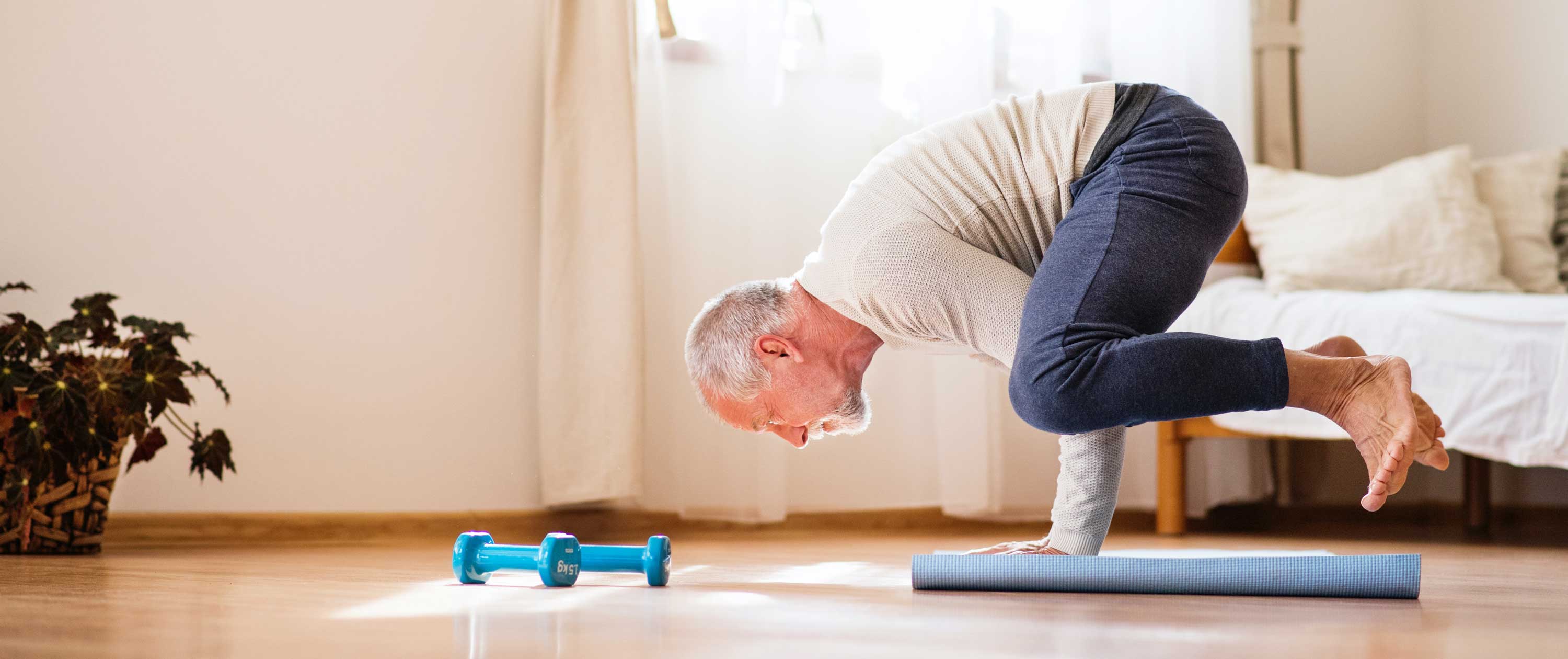 Elderly man practicing yoga on mat in bright room, balancing in crow pose. Blue dumbbells nearby. Atmosphere is calm and focused.