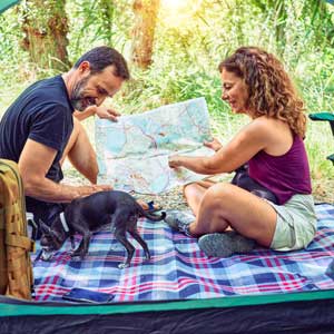 A couple sits on a plaid blanket inside a tent, looking at a map. A small black dog plays nearby. The scene is set outdoors, conveying a joyful camping vibe.