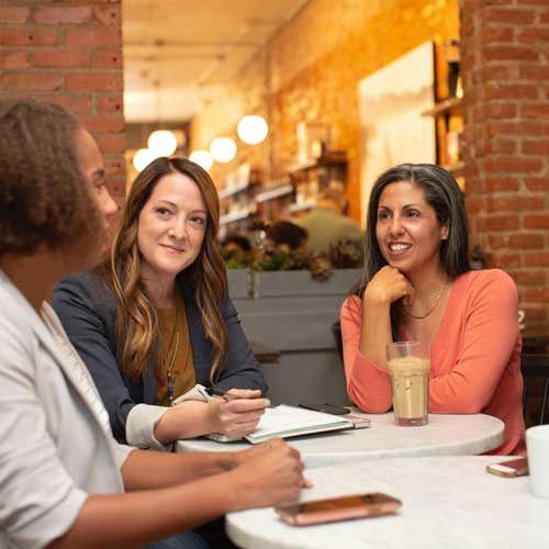 Three women sit at a round cafe table, smiling and engaged in conversation. The warm, cosy setting features brick walls and soft lighting.